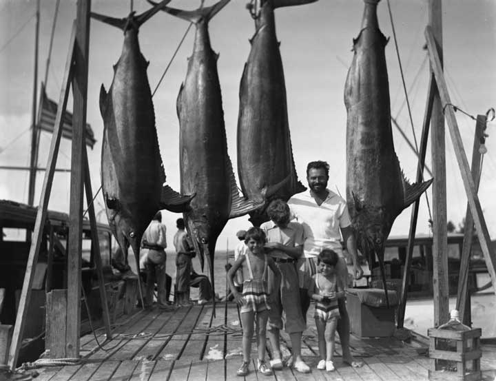 Black and white photo of Ernest Hemingway with his three sons on a dock in front of four large hanging sword fish.