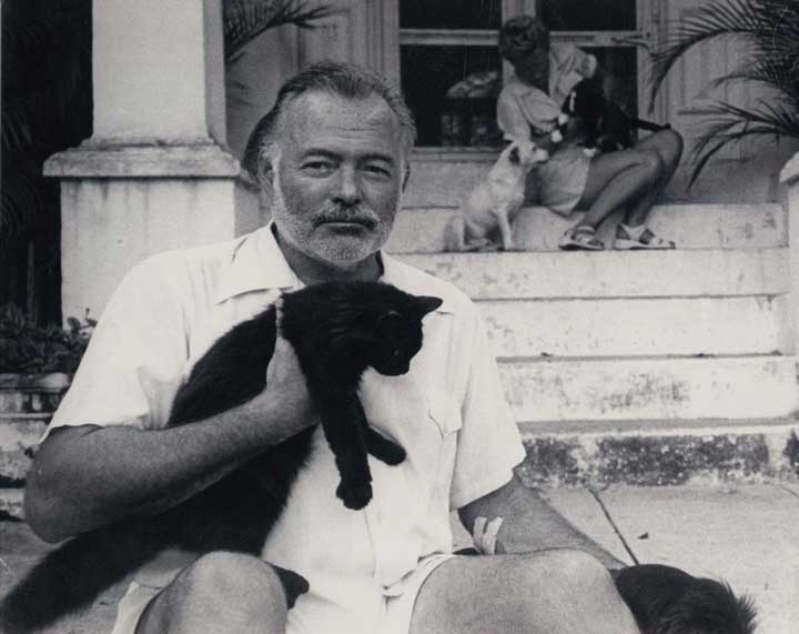 Black and white photo of Ernest Hemingway sitting on a porch holding a black cat.