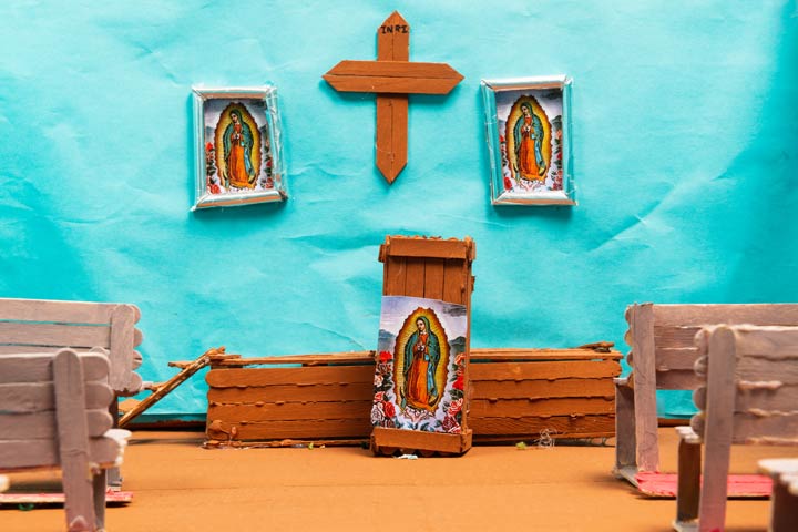 Diorama of a church scene. The furniture and a crucifix are made of popsicle sticks. There are three images of the Virgin Mary. The wall is made of blue construction paper.