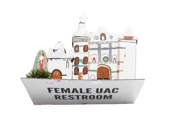 Hand-painted cardboard sculpture of a church with a Virgin Mary outside. The bottom of the cardboard sculpture says "FEMALE UAC RESTROOM" in bold black letters