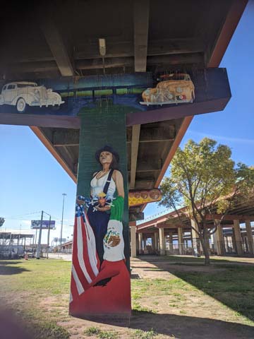 Mural under an overpass. Depicts a woman standing and holding a Mexican flag and an American flag. Above her is a painting of the overpass and two vintage cars.