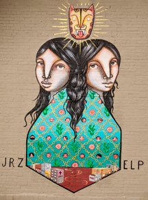 Mural of two girls back to back. They have black hair. A jaguar head floats above them. They are wearing a blue green dress with a chain pattern containing flowers, cacti, and human faces. At the bottom are two cityscapes, Juarez on the left and El Paso on the right. The landscape is brown dirt.