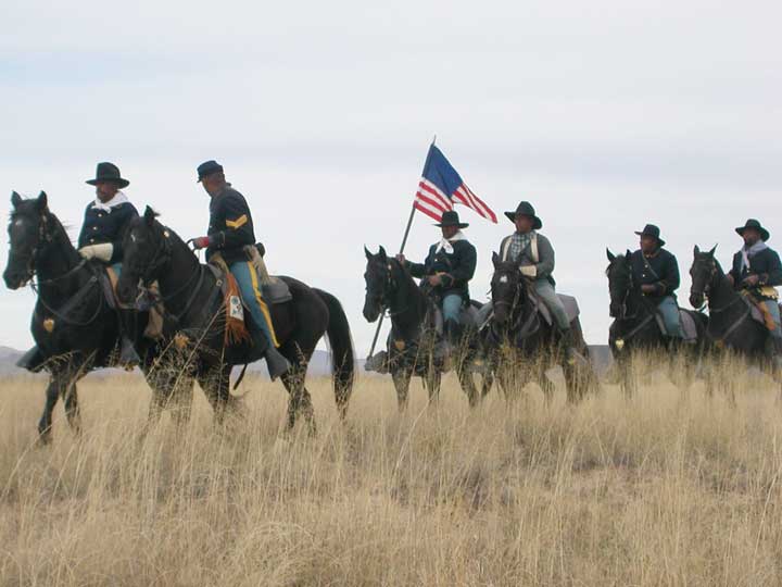 A photo of a reenactment of Buffalo Soldiers at Fort Craig. 6 men in period uniforms are riding horses through a field. The man in the middle of the photo holds an American flag.
