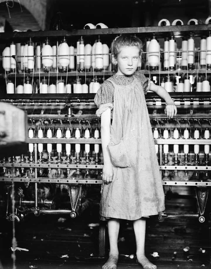 Black and white photo of a 12 year old girl standing in front of factory equipment. Her dress is dirty and she has bare feet.