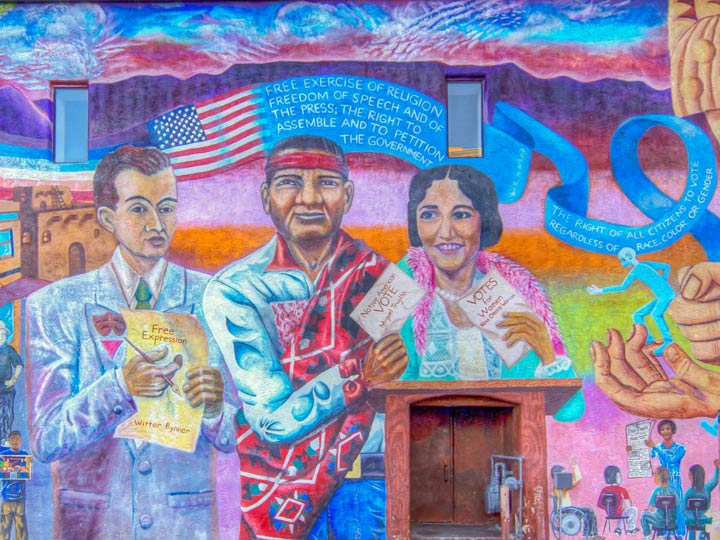 Brightly colored mural of Witter Bynner, Miguel Trujillo Sr., & Nina Otero Warren standing in front of NM mountain range. A banner in the back has an American flag and text that reads "FREE EXERCISE OF RELIGION FREEDOM OF SPEECH AND OF THE PRESS; THE RIGHT TO ASSEMBLE AND TO PETITION THE GOVERNMENT... THE RIGHT OF ALL CITIZENS TO VOTE REGARDLESS OF RACE, COLOR OR GENDER"