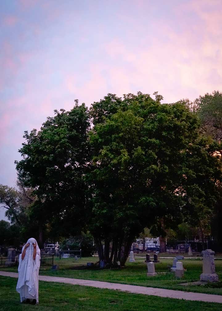 Photo of a person in a ghost costume crying in a cemetery at sunset.