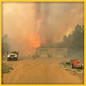 Photo of a wildfire burning tall trees behind a dirt lot and garage. The sky is filled with smoke and tinted yellow.