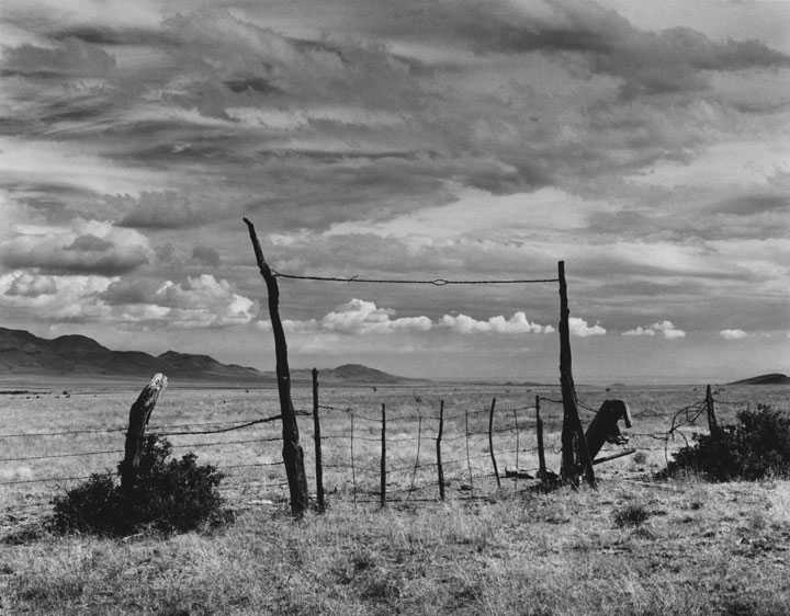 Black and white photo of a wire and wooden fence in an empty field of dirt and sagebrush. Hills in the background.