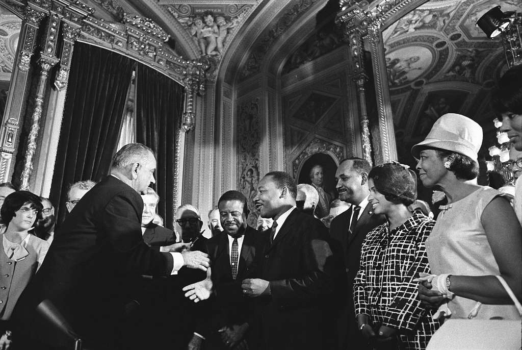 Black and white photo of Dr. Martin Luther King Jr. shaking hands with President Lyndon B. Johnson in a crowd of people.