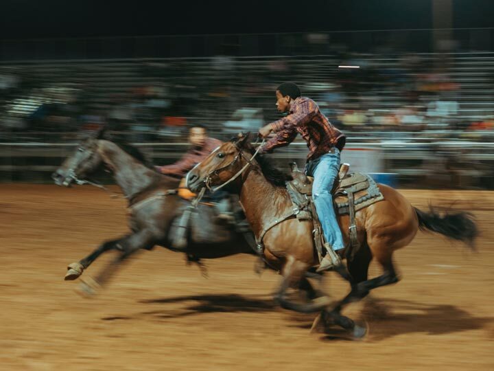 Photo of two men riding running horses in a stadium. There is a fast motion blur.