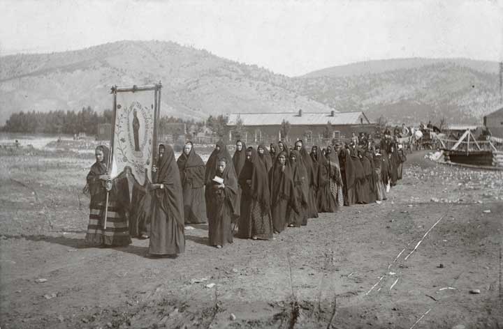 Black and white photo of a funeral procession. Everyone is wearing black shawls/tápalos and the people at the front of the procession are holding a banner with an image of a saint