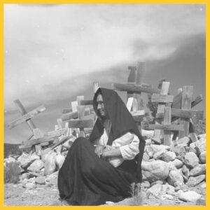 Black and white photo of a woman in a black shawl/tápalo in front of a mound of rocks and crucifixes
