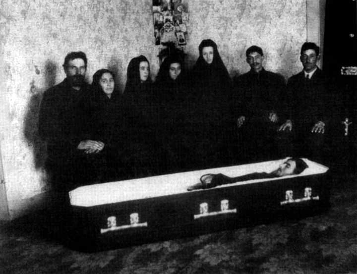 Black and white photo of 7 people standing behind an open casket, mourning a family member