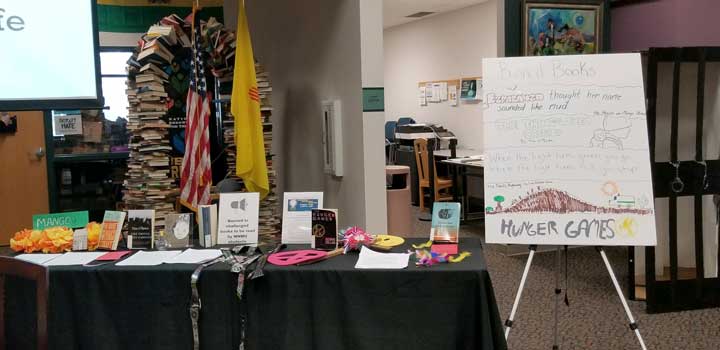 A photo of a banned book table display. The table features various banned books, an arch made of books and the US and NM flags are in the background, a handmade "Banned Books" poster is on the right listing titles of banned books