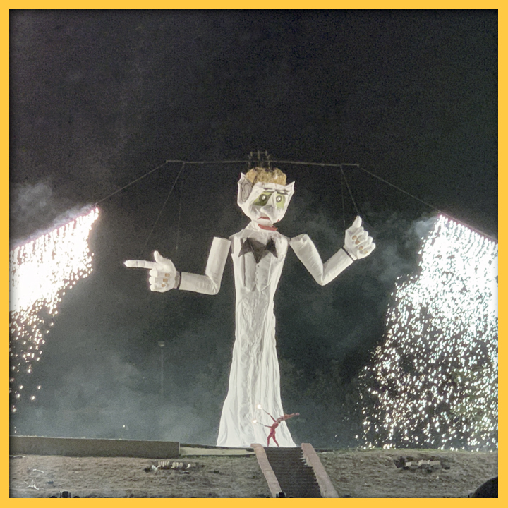Zozobra, a 50 foot effigy of "old man gloom," with a person dressed in red as the fire spirit in front of it. Waterfalls of fire on either side.