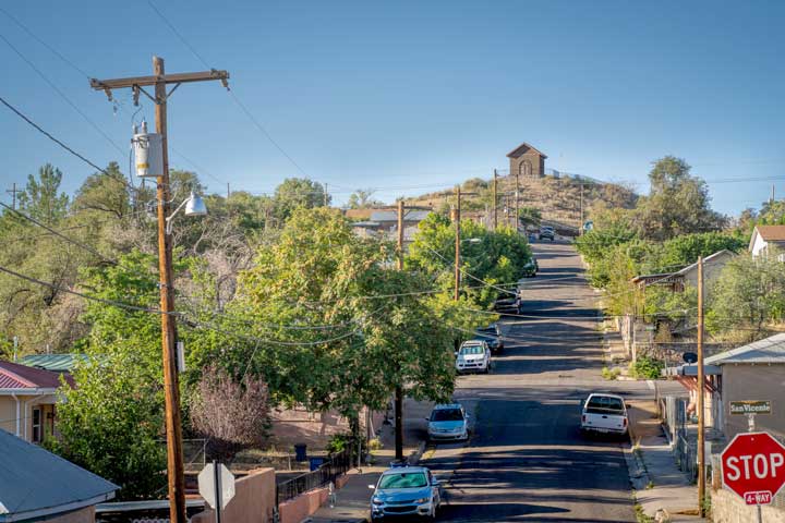 A photo of a road going up Chihuahua Hill. The street is lined with trees and parked cars. There is a small building at the top of the dirt hill at the end of the road.