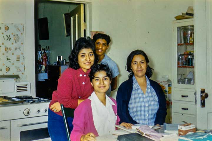 A photo of two women at a kitchen table with books, a woman and a boy are standing behind them. Everyone is smiling at the camera.