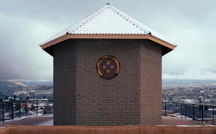 A photo of a small hexagonal brick building feature with a tin roof. There is a grated metal circle in the middle of the wall with a Zia symbol.