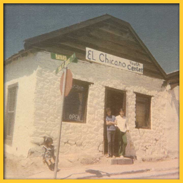 A photo of a white stone building with a hand-painted sign above the door that reads "El Chicano Youth Center." Two people are standing in the doorway.