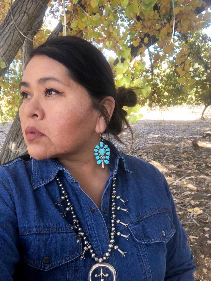 A photo portrait of Venya Yazzie wearing turquoise earrings and standing outside under a tree
