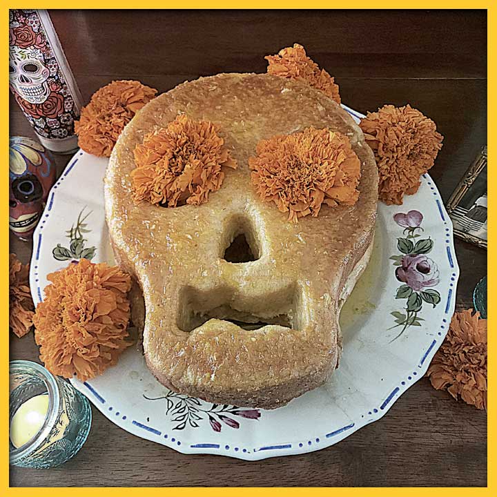 Pan de Muerto, skull shaped bread with orange marigold flowers on a white flowered plate on an altar table