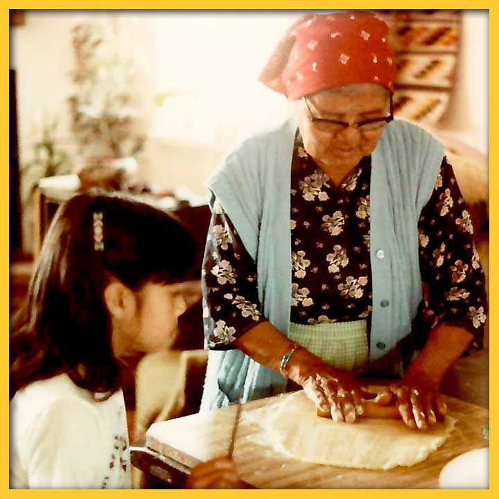 Photo of a woman in a red bandana rolling out cookie dough while her granddaughter watches