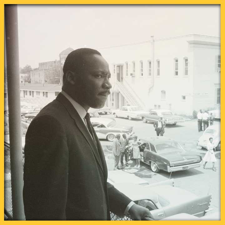 Photo of Martin Luther King, Jr. standing on a balcony overlooking a parking lot.